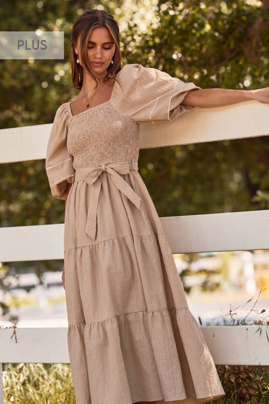 Woman wearing a beige dress with puffed sleeves standing by a white fence outdoors.